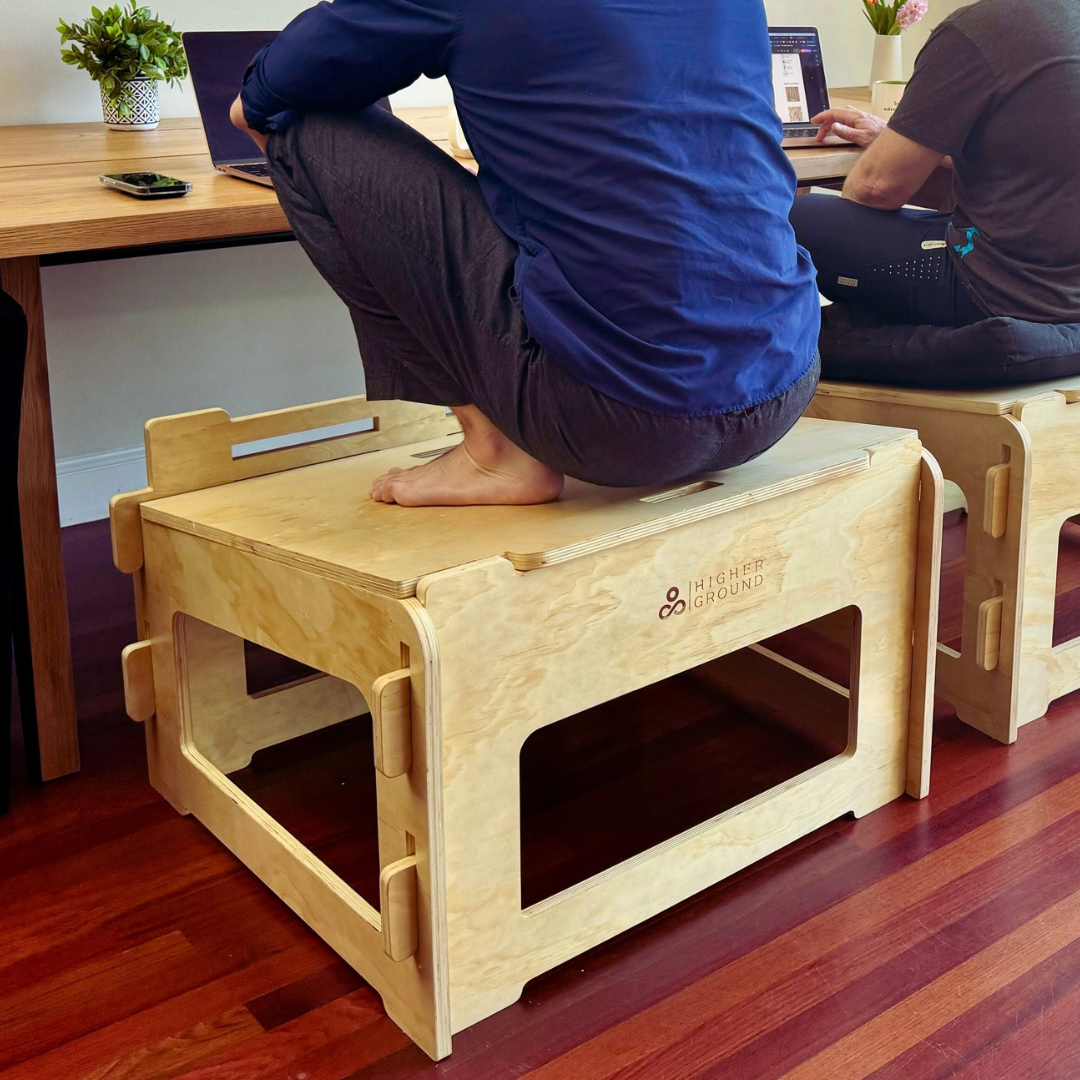 A man squats on a wooden Higher Ground Chair at a desk