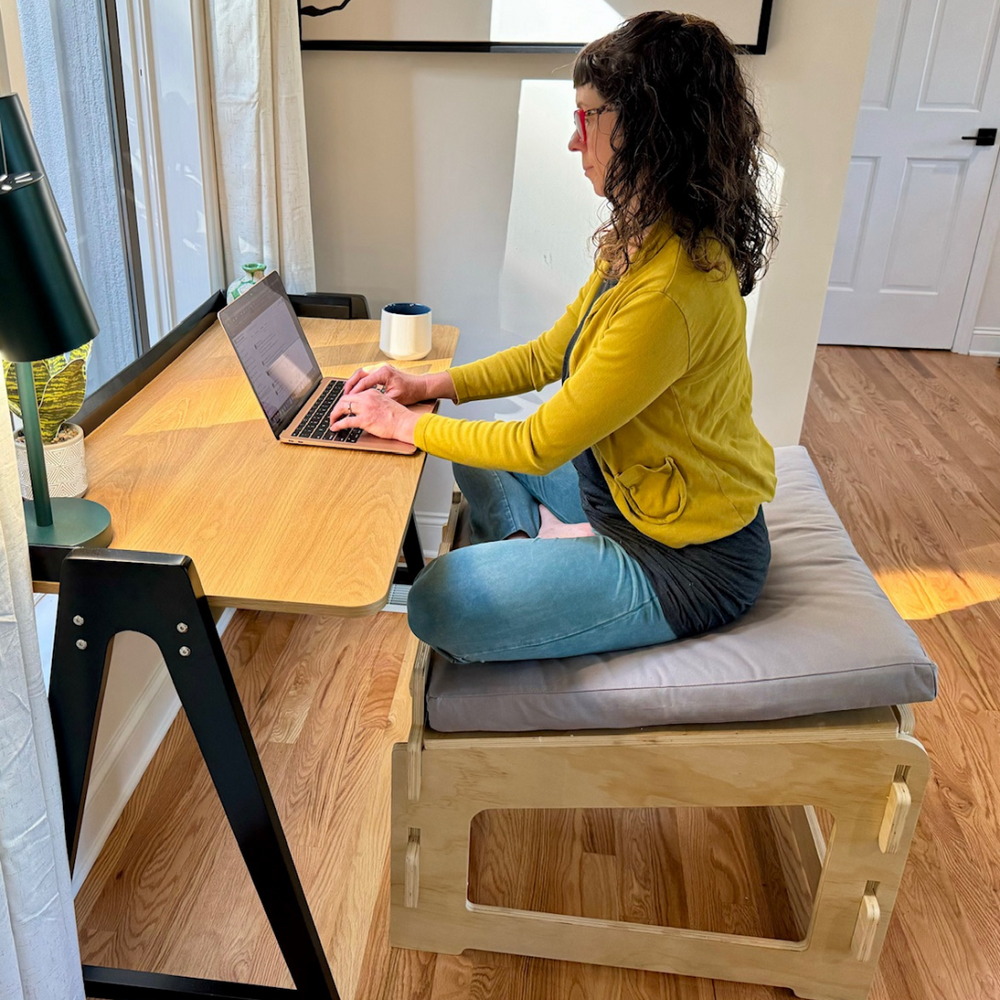 
                      
                        Woman sitting on chair with a grey cushion, in front of a wooden desk ,and typing on a laptop 
                      
                    
